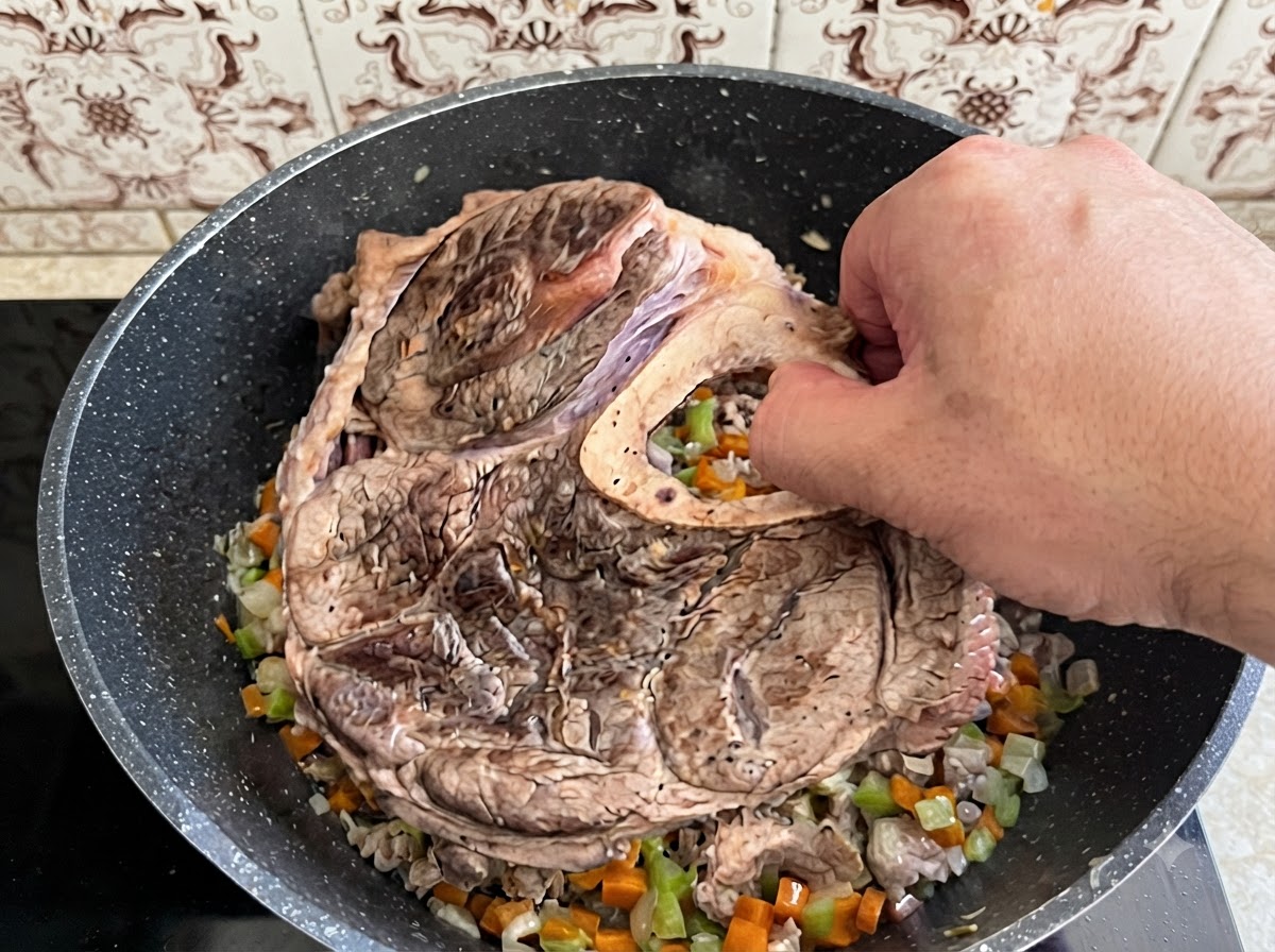 Una mano colocando un gran trozo de jarrete de ternera sellado y hueso en una olla llena de verduras picadas cocidas.
