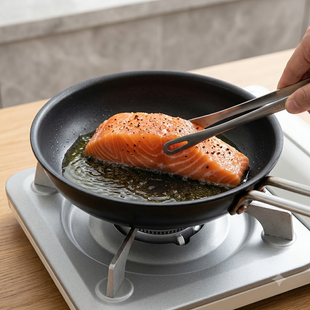 Un filete de salmón fresco siendo colocado con la piel hacia abajo en una sartén caliente con aceite usando pinzas de cocina.