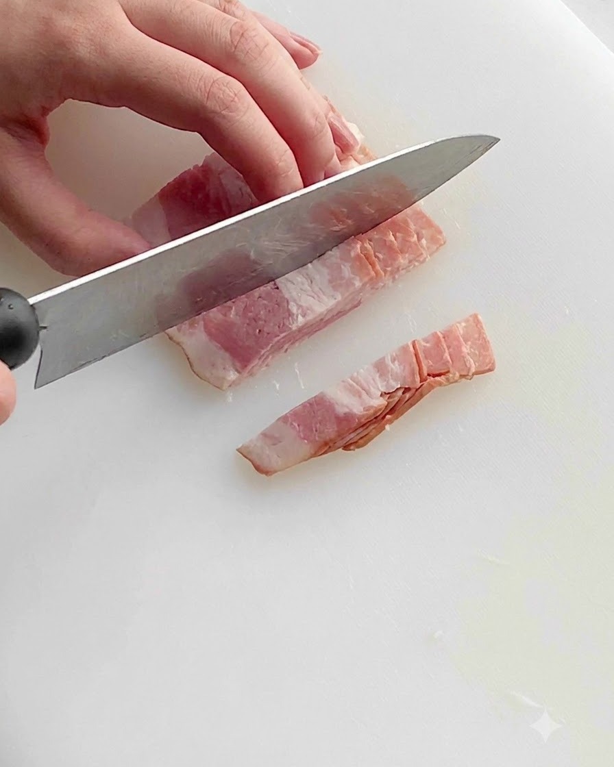 Un cuchillo de cocina cortando tiras de tocino crudo en trozos pequeños y uniformes sobre una tabla de cortar de plástico blanco.