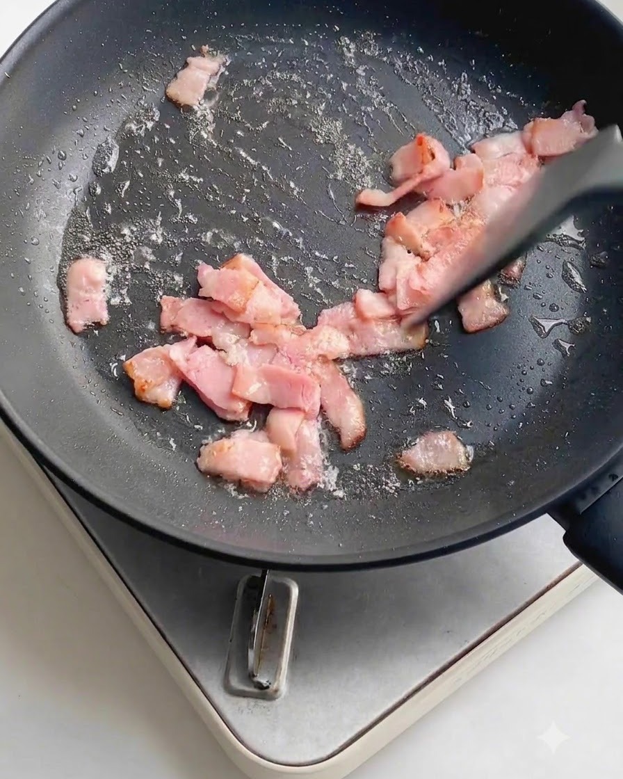 Ajo fresco picado finamente esparcido sobre los trozos de tocino frito dentro de una sartén antiadherente oscura.