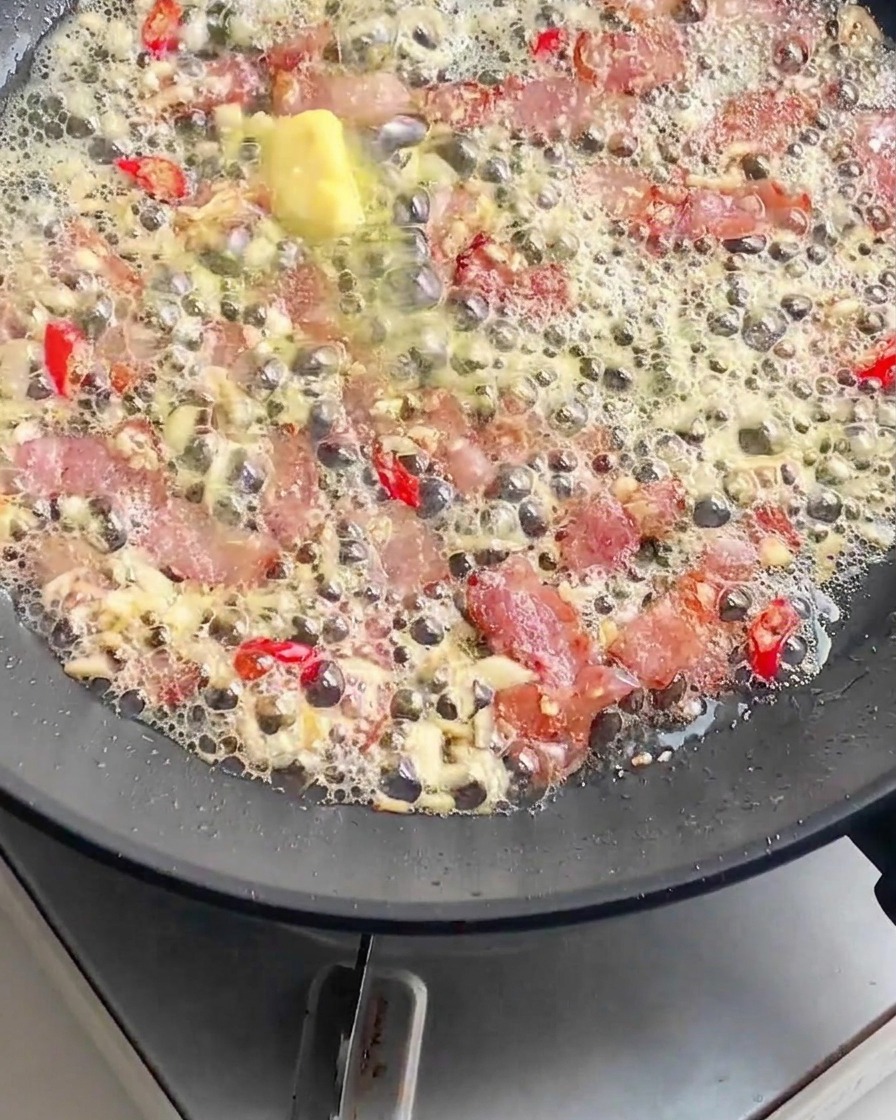 Fideos de pasta cocidos siendo añadidos a una sartén llena de tocino burbujeante, mantequilla derretida, ajo picado y chiles rojos en rodajas.