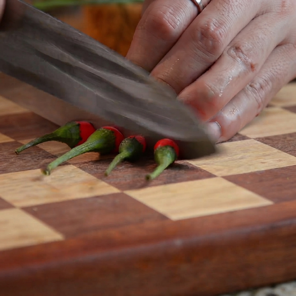 Un cuchillo de cocina cortando chiles rojos en trozos pequeños sobre una tabla de cortar de madera.