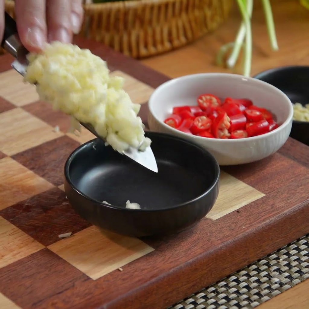 Ajo picado en la hoja de un cuchillo siendo transferido a un bol negro junto con un bol de chiles rojos picados.