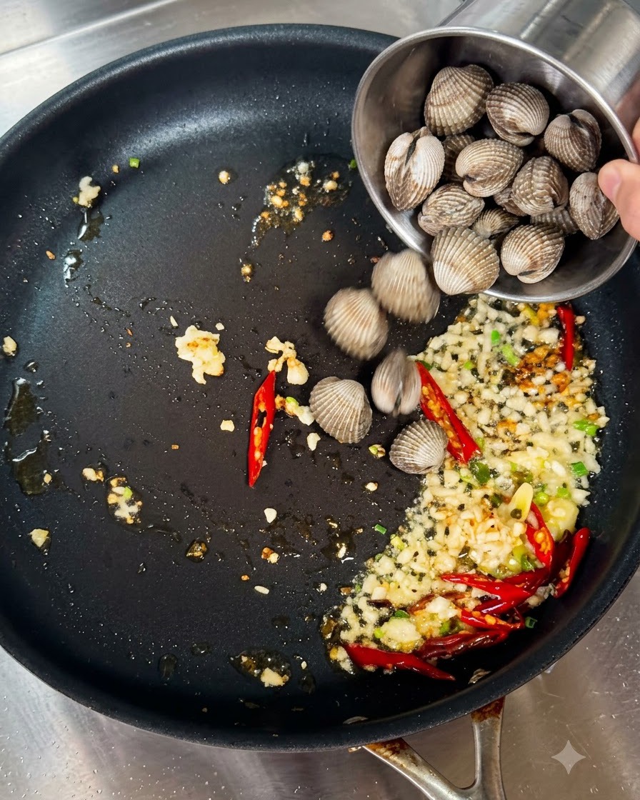 Almejas frescas siendo vertidas de una taza de metal pequeña en una sartén con ajo y chiles salteados.