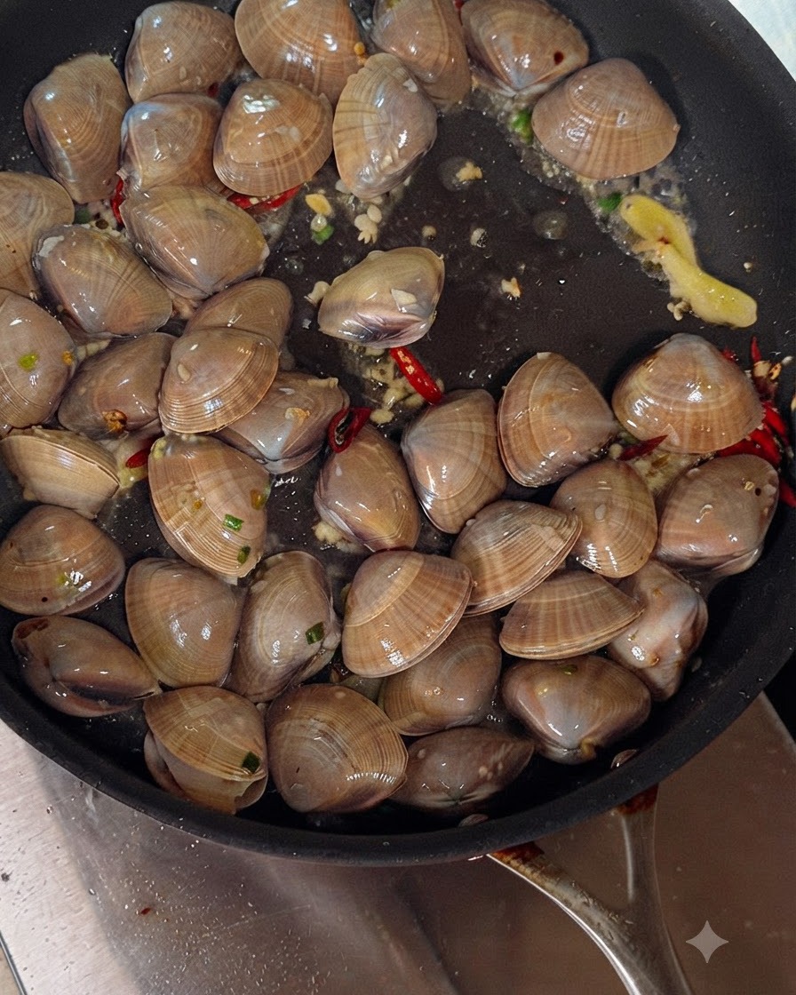 Vino blanco siendo vertido de una botella en una sartén chisporroteante llena de almejas, ajo y chiles.