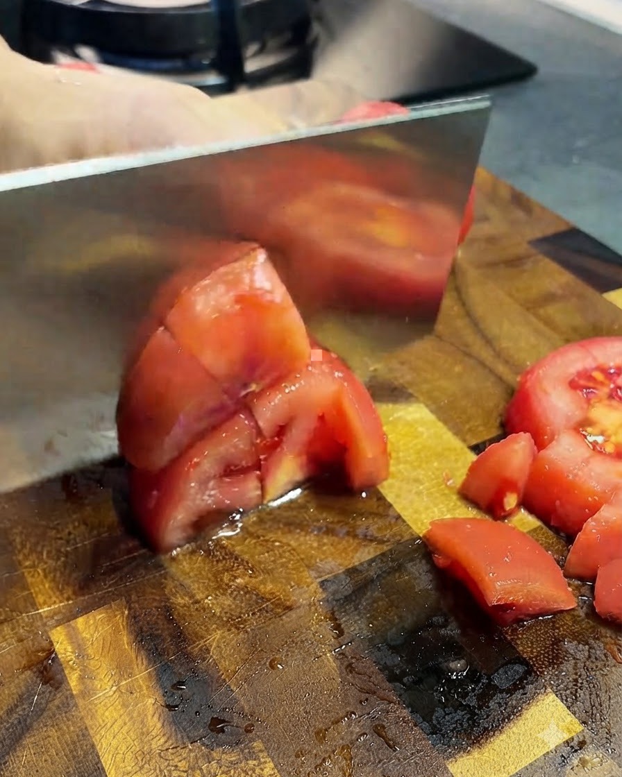 Tomates pelados siendo cortados en trozos pequeños con un cuchillo de cocina sobre una tabla de madera.