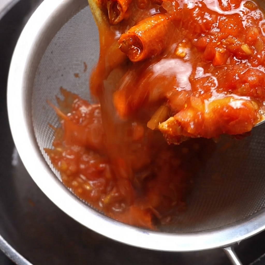 Salsa de tomate cocida siendo vertida a través de un colador de metal con una espátula presionando el contenido.