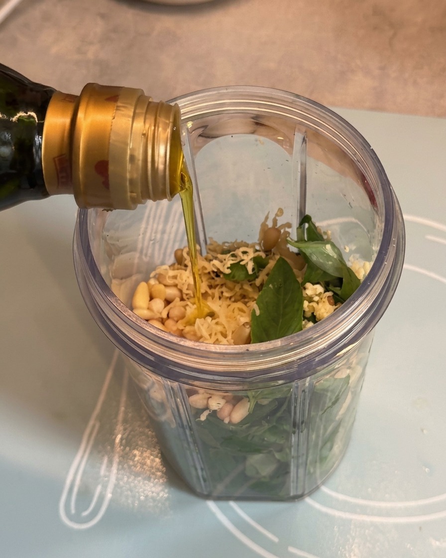 Aceite de oliva cayendo desde una botella hacia el vaso de la licuadora sobre los ingredientes del pesto.