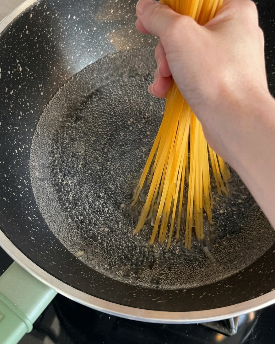 Fideos espagueti secos sostenidos por una mano y colocados dentro de una olla con agua hirviendo.