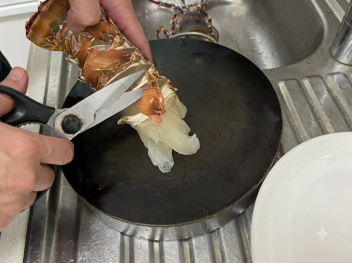 Manos usando tijeras de cocina para cortar a lo largo de la membrana inferior de una cola de langosta cruda sobre una superficie de corte oscura.
