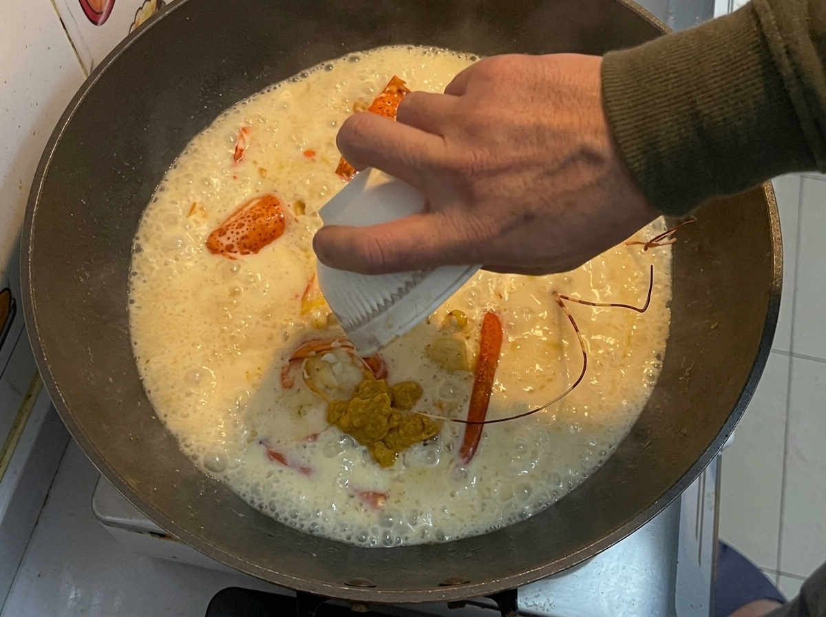 La pasta amarilla de langosta reservada se vierte desde un pequeño tazón blanco en un wok con trozos de langosta frita y una salsa cremosa.
