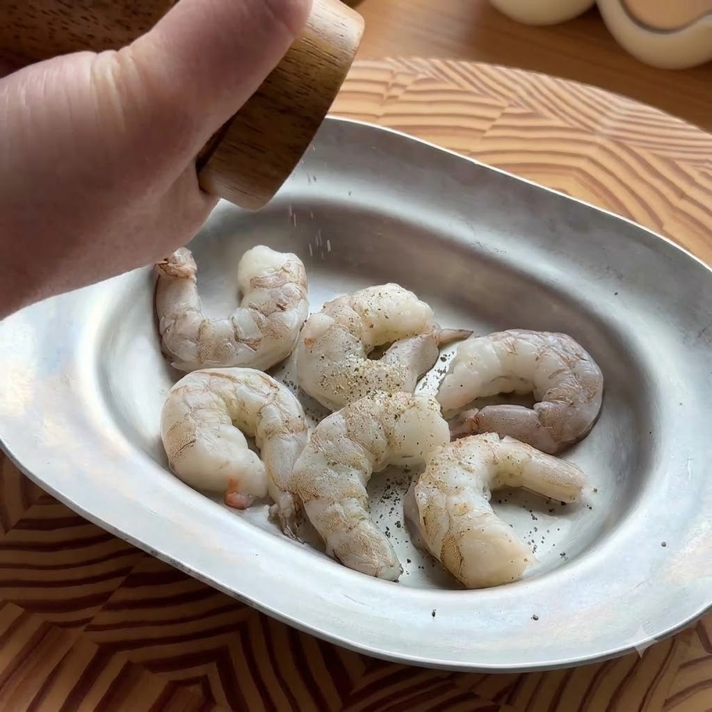 Una mano usando un molinillo de pimienta de madera para sazonar camarones crudos y pelados en una bandeja de metal ovalada.
