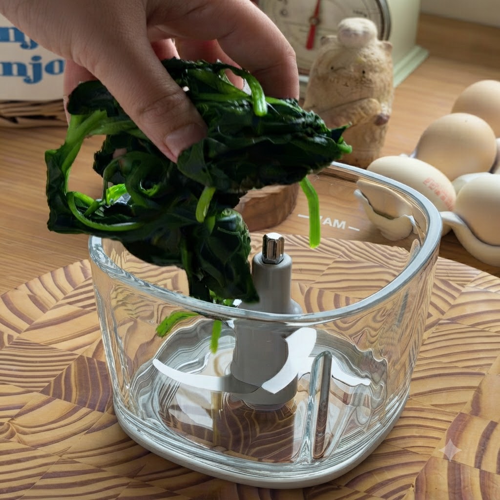 Una mano colocando un puñado de espinaca cocida y escurrida en el vaso vacío de un procesador de alimentos de vidrio.