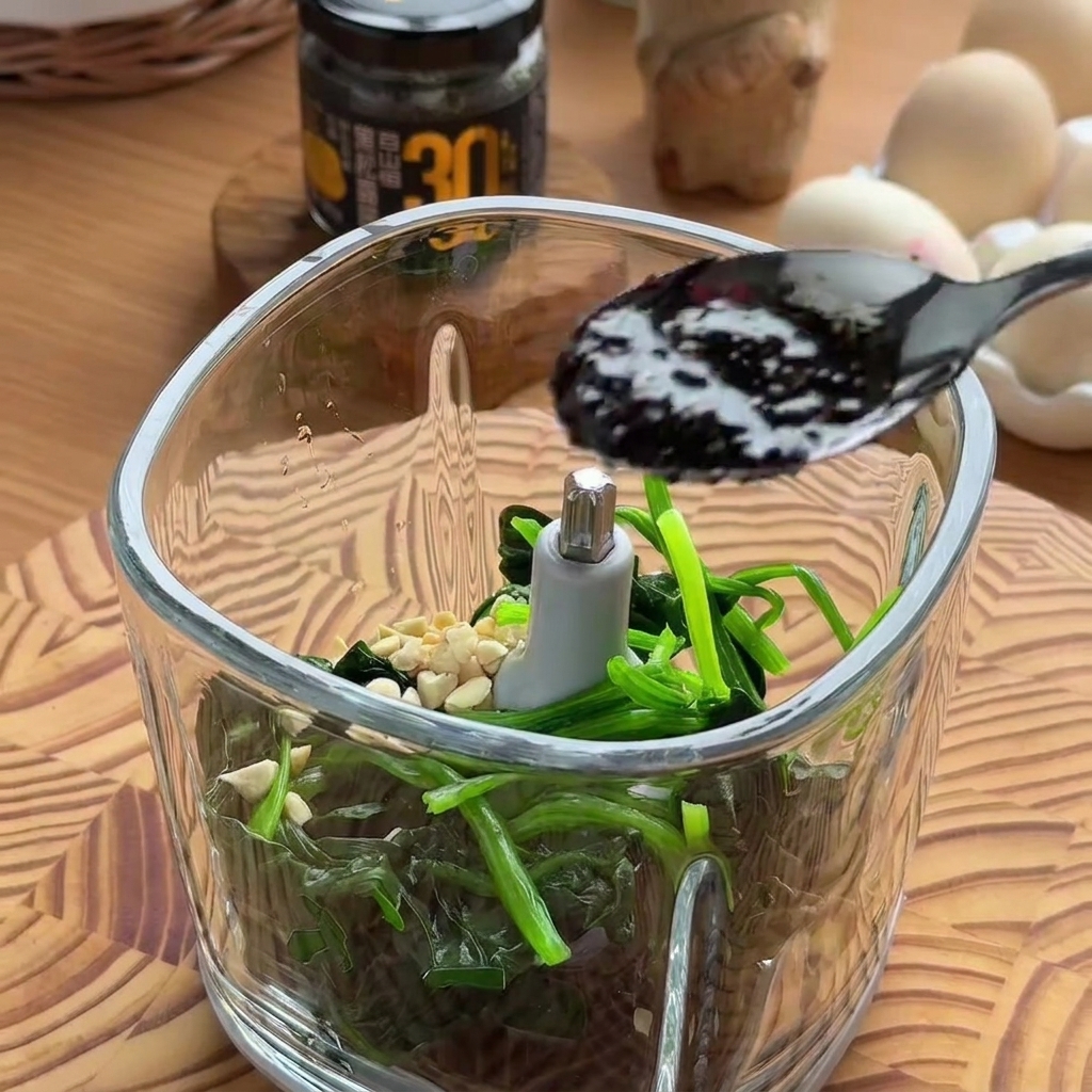 Una cuchara negra dejando caer salsa de trufa negra oscura y rica en el vaso de un procesador de alimentos lleno de espinacas y frutos secos.