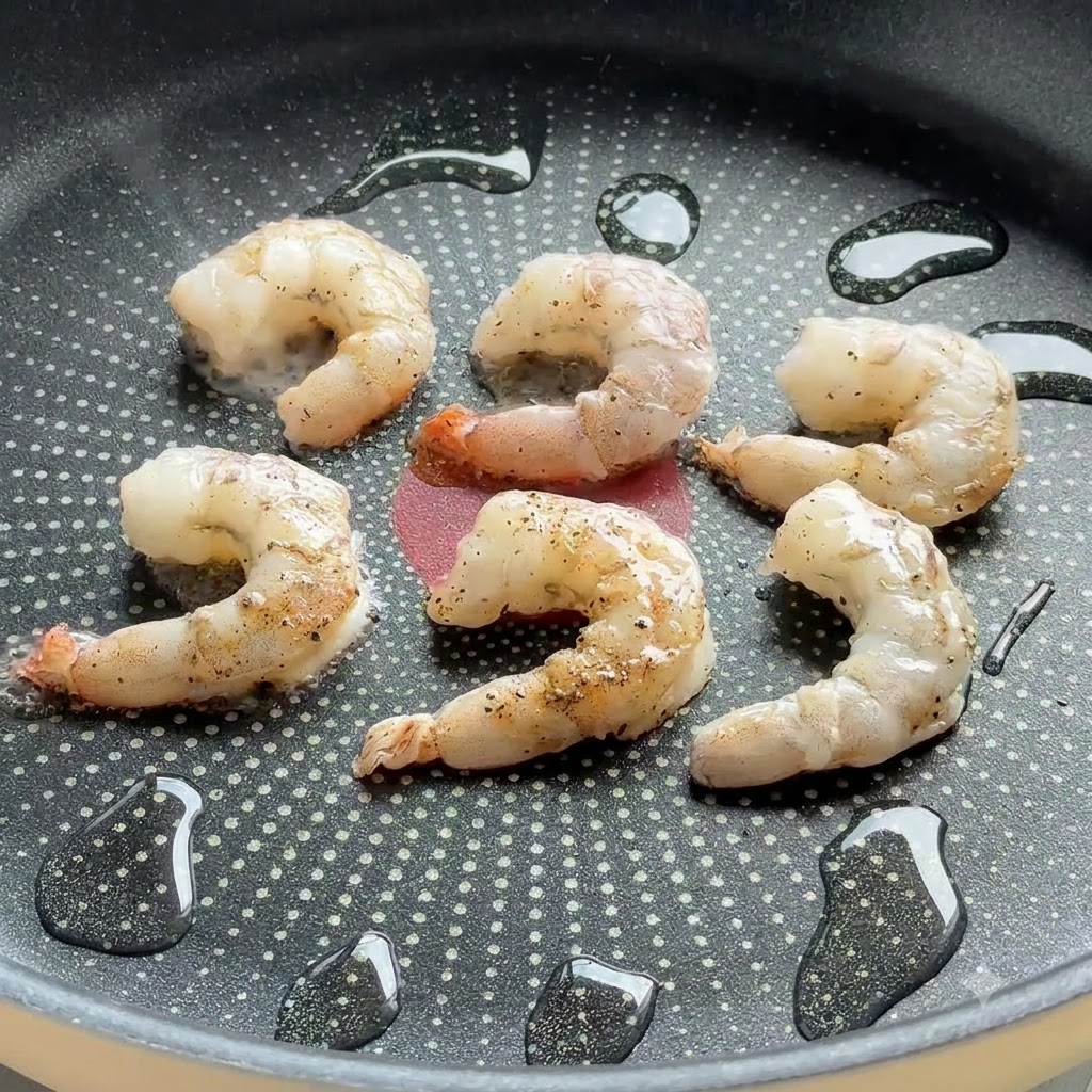Camarones sazonados chisporroteando en una sartén antiadherente con gotas de aceite.
