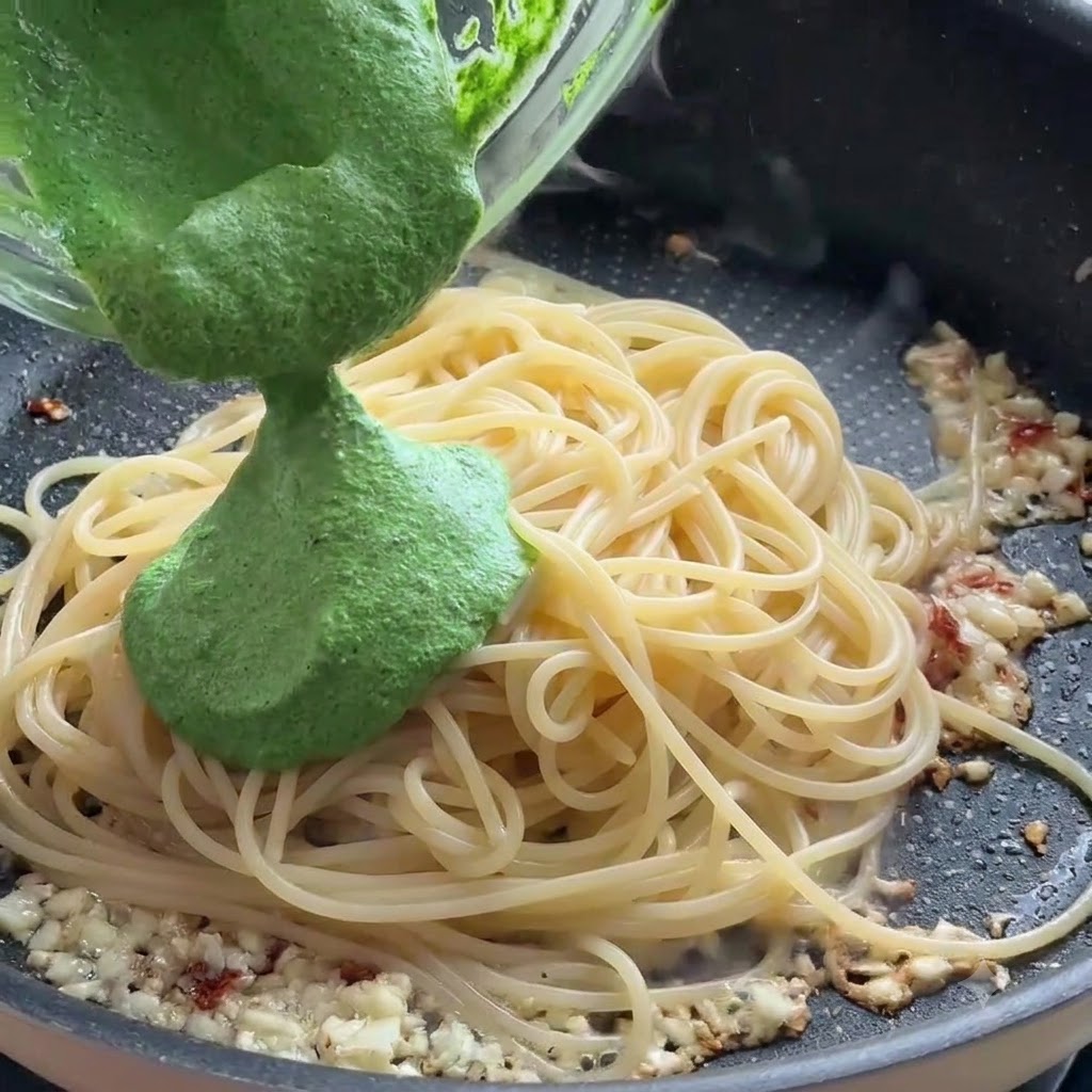 Salsa de pesto verde siendo vertida desde un bol de vidrio sobre los fideos de espagueti en una sartén.