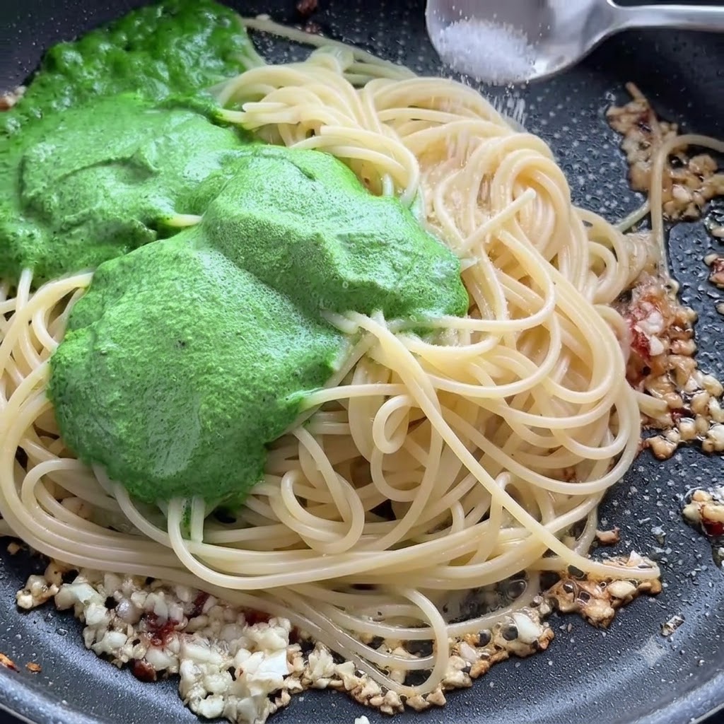 Una cucharada de sal siendo espolvoreada sobre los fideos de espagueti y la salsa de pesto verde en una sartén.