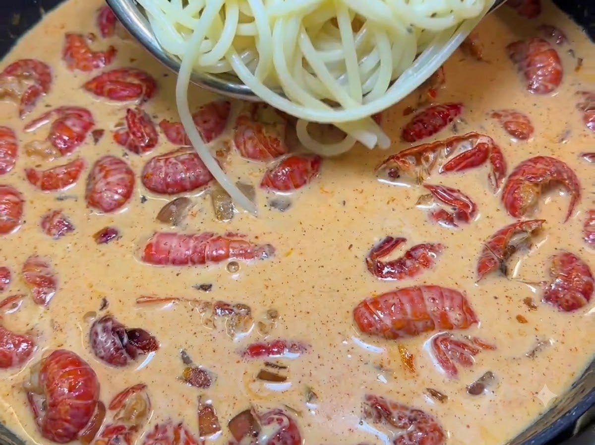 Pinzas levantando fideos de espagueti cocidos hacia una sartén chispeante llena de salsa cremosa rosada y colas de cangrejo de río rojas.