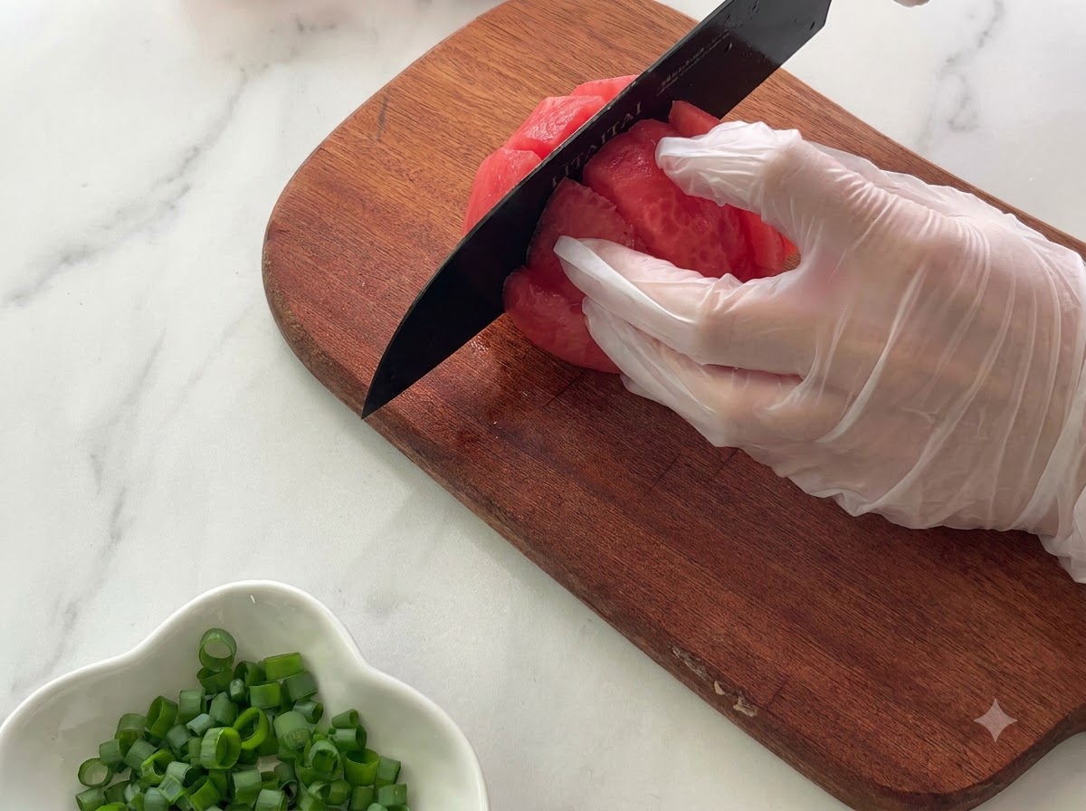 Una mano con guantes sosteniendo un tomate en una tabla de cortar de madera mientras lo corta en trozos con un cuchillo negro.
