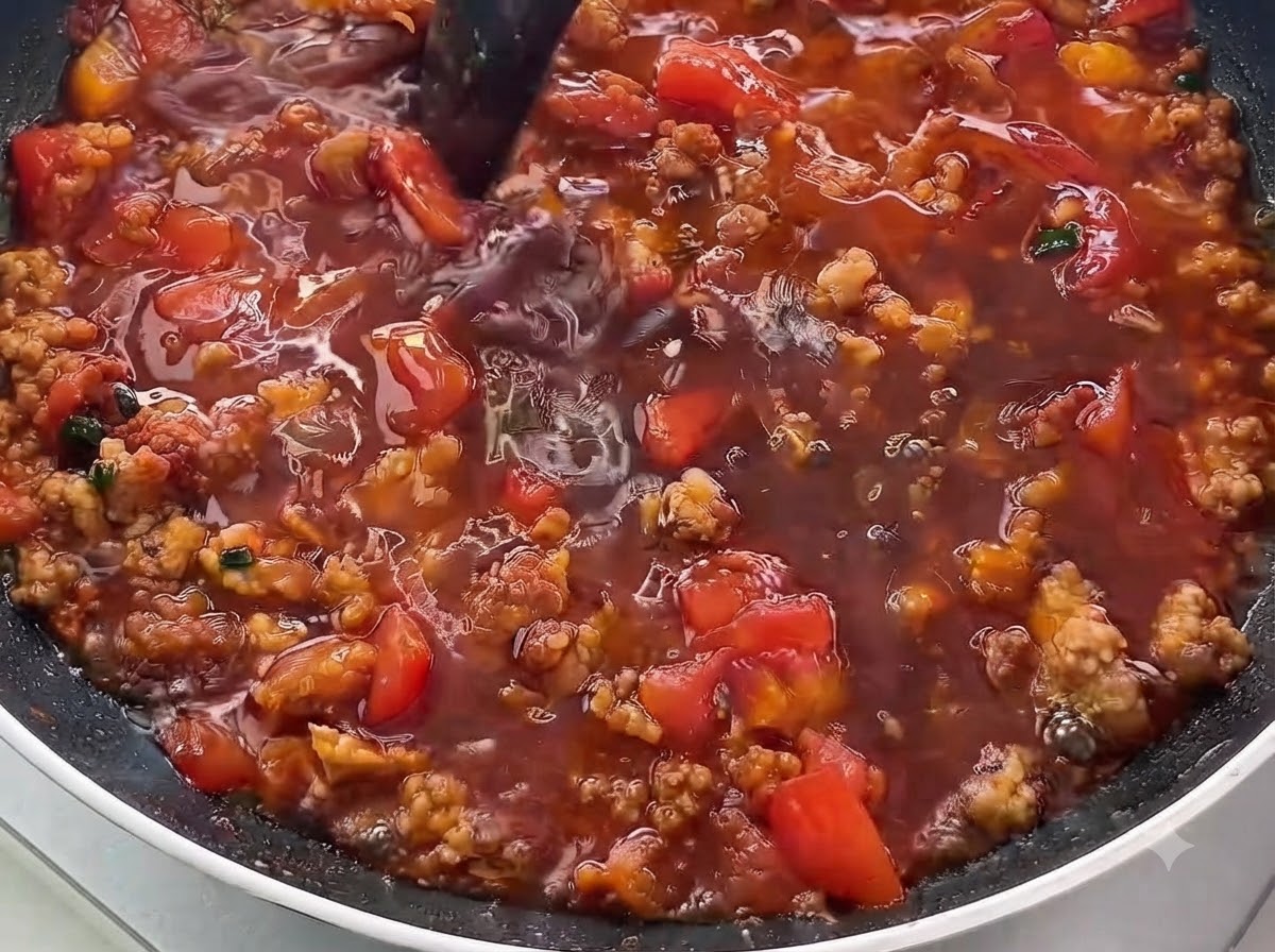 Un primer plano de salsa líquida oscura siendo vertida en una sartén llena de carne picada dorada y tomates en dados.