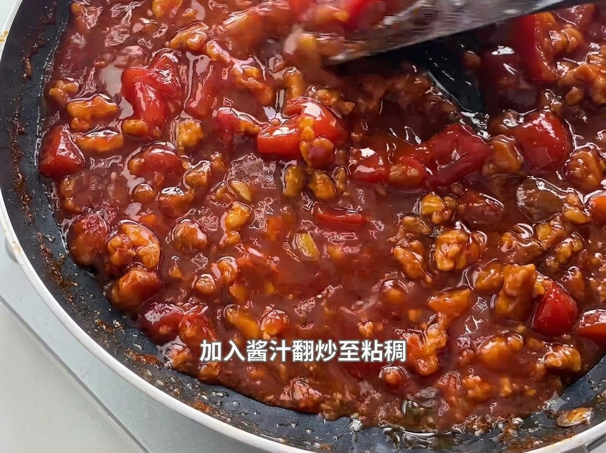Una espátula removiendo una salsa espesa y burbujeante de tomate y carne picada cocinándose en una sartén negra.