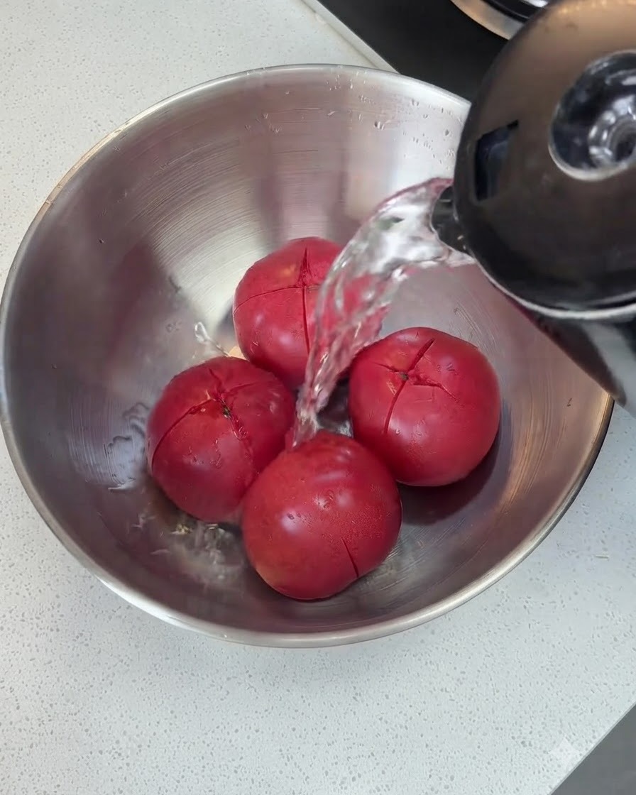 Agua caliente cayendo desde una tetera sobre los tomates con incisiones en un tazón de metal.