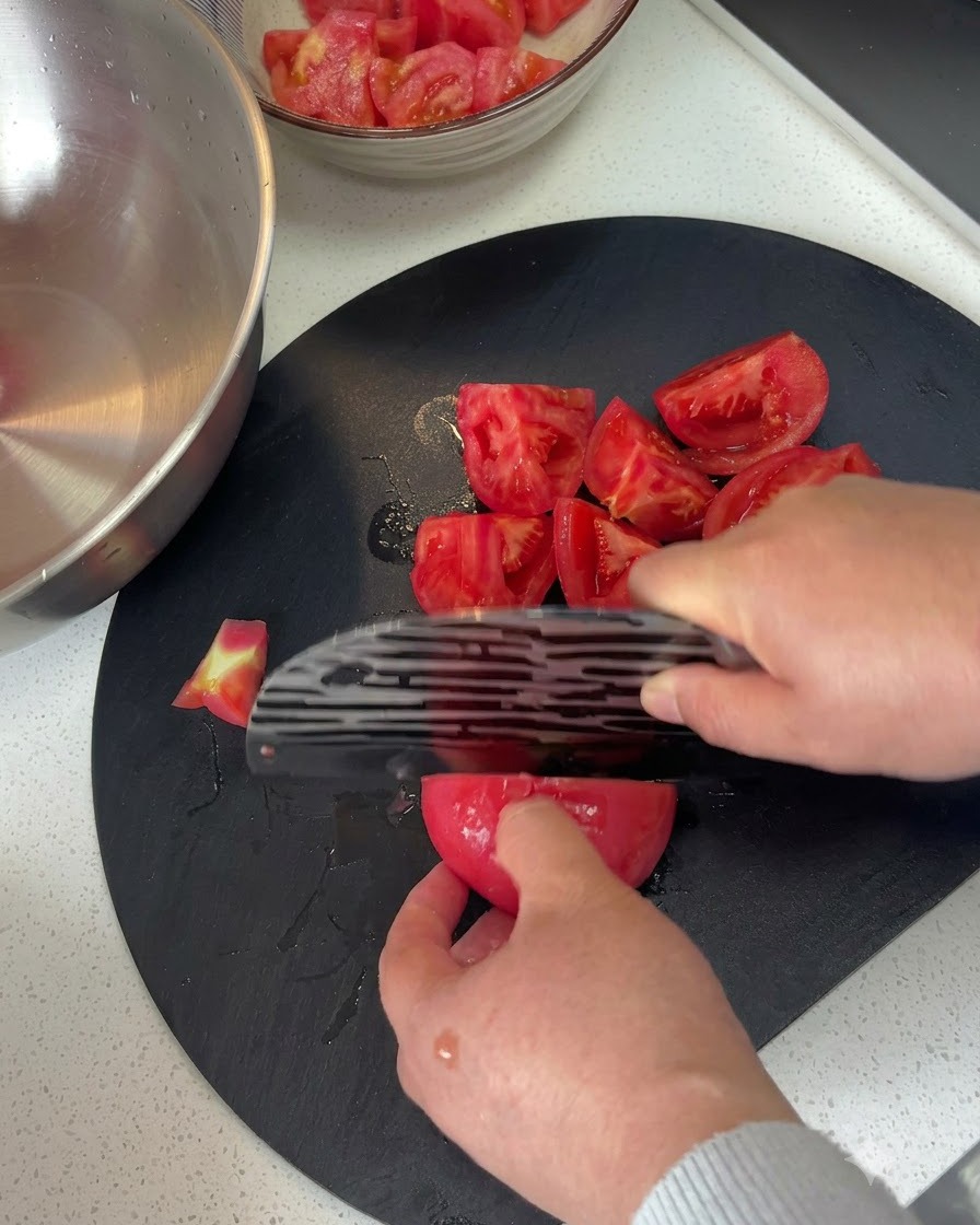 Tomates pelados siendo cortados en trozos sobre una tabla de cortar oscura con un cuchillo.