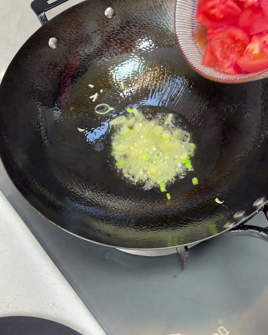 Trozos de tomate siendo vertidos desde un tazón hacia un wok con cebolletas salteadas y aceite.