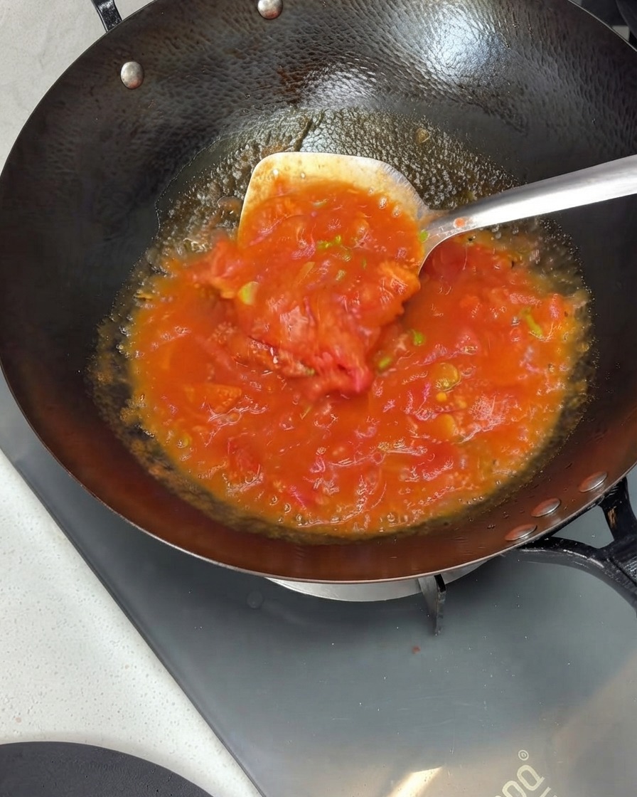 Tomates siendo machacados y revueltos en una salsa rica dentro de un wok usando una espátula de metal.