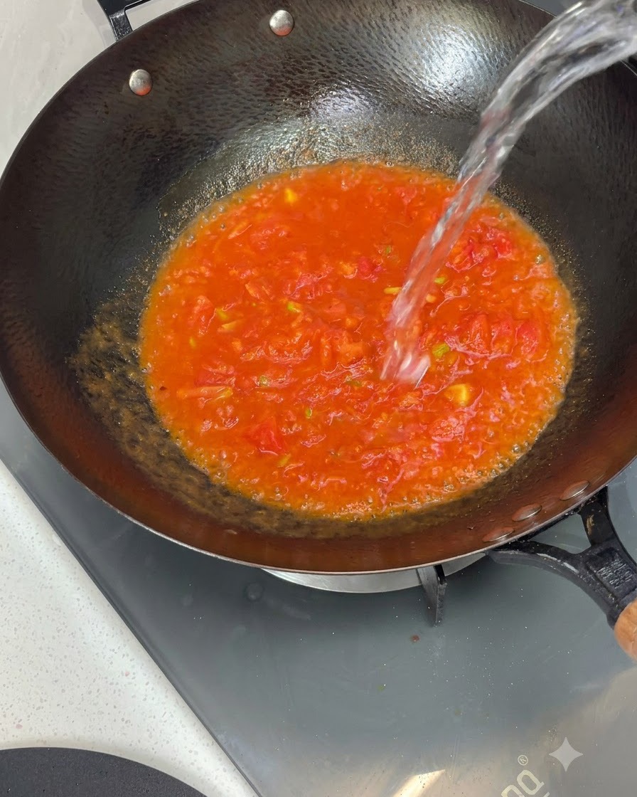 Agua siendo vertida desde arriba en un wok lleno de salsa de tomate cocida.