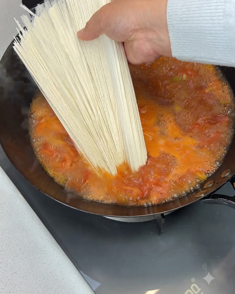 Un paquete de fideos secos sostenido sobre y añadido a una sopa de tomate burbujeante en un wok.