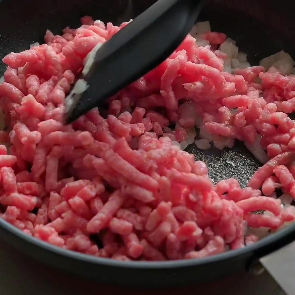 Carne picada de res rosada cruda siendo desmenuzada y salteada con cebollas en una sartén negra.