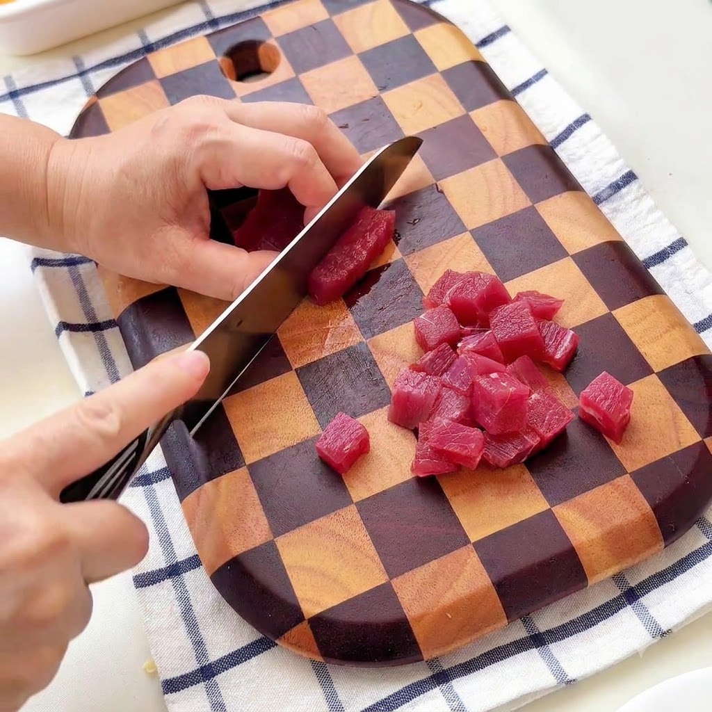 Una persona cortando carne de res en cubos pequeños sobre una tabla de cortar de madera a cuadros.