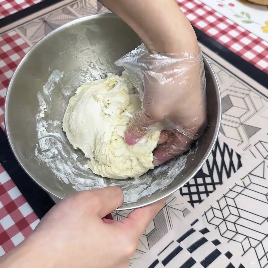 Una mano con guante amasando masa húmeda de pizza dentro de un bol de acero inoxidable.