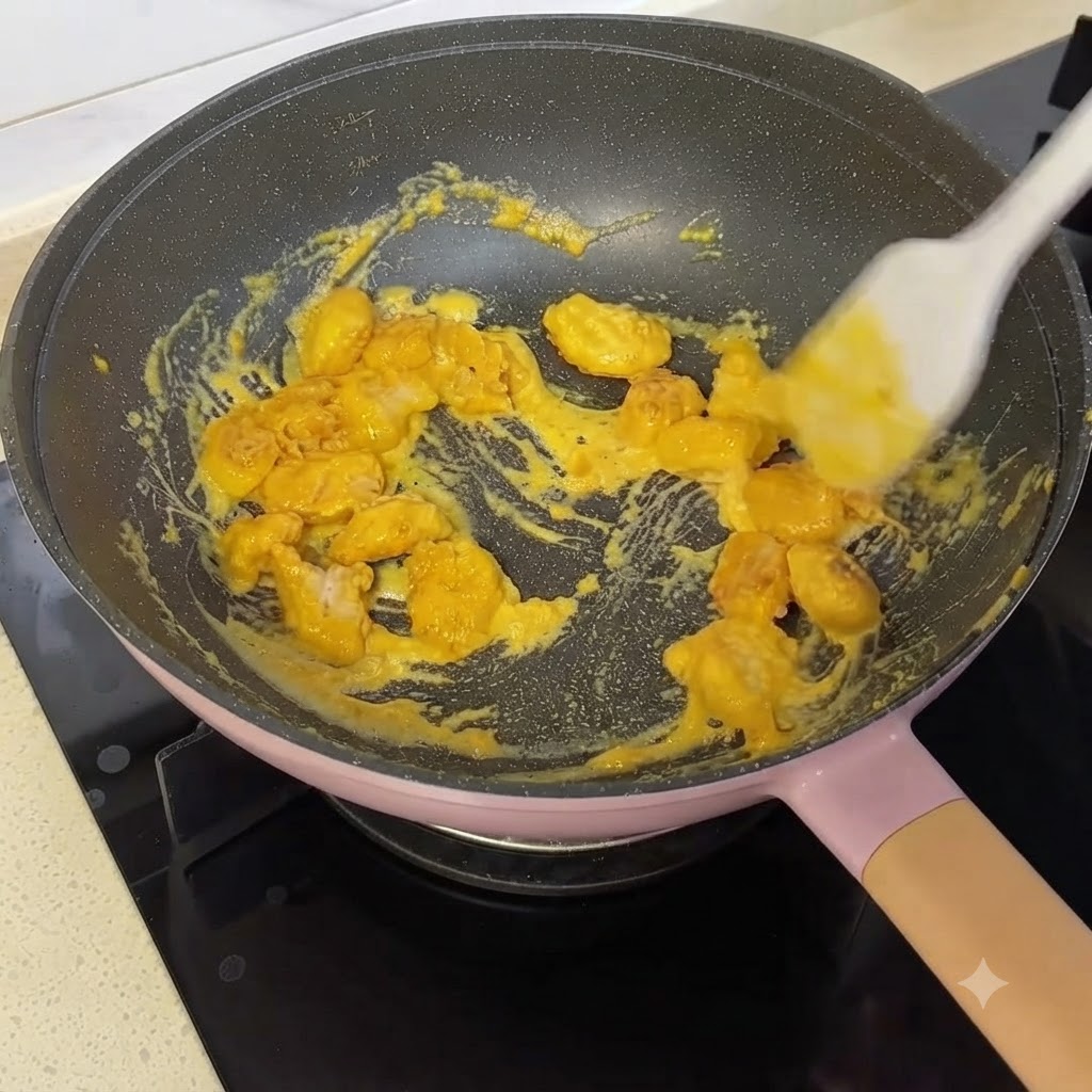 Trozos de pollo crujiente siendo removidos y cubiertos con salsa amarilla de yema de huevo salada en una sartén usando una espátula de silicona.