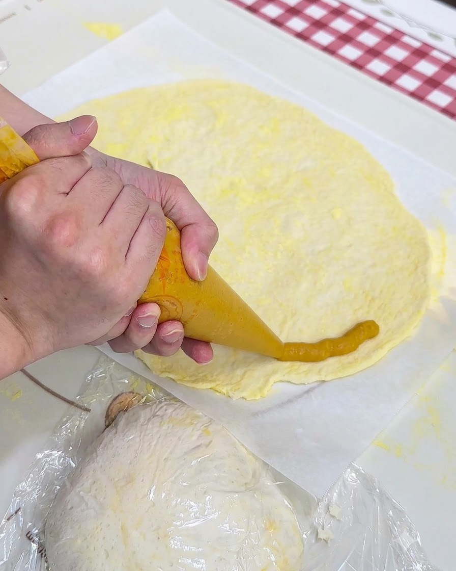 Un par de manos usando una manga pastelera para aplicar puré de batata amarillo alrededor del borde de una lámina de masa de pizza plana sobre papel pergamino.