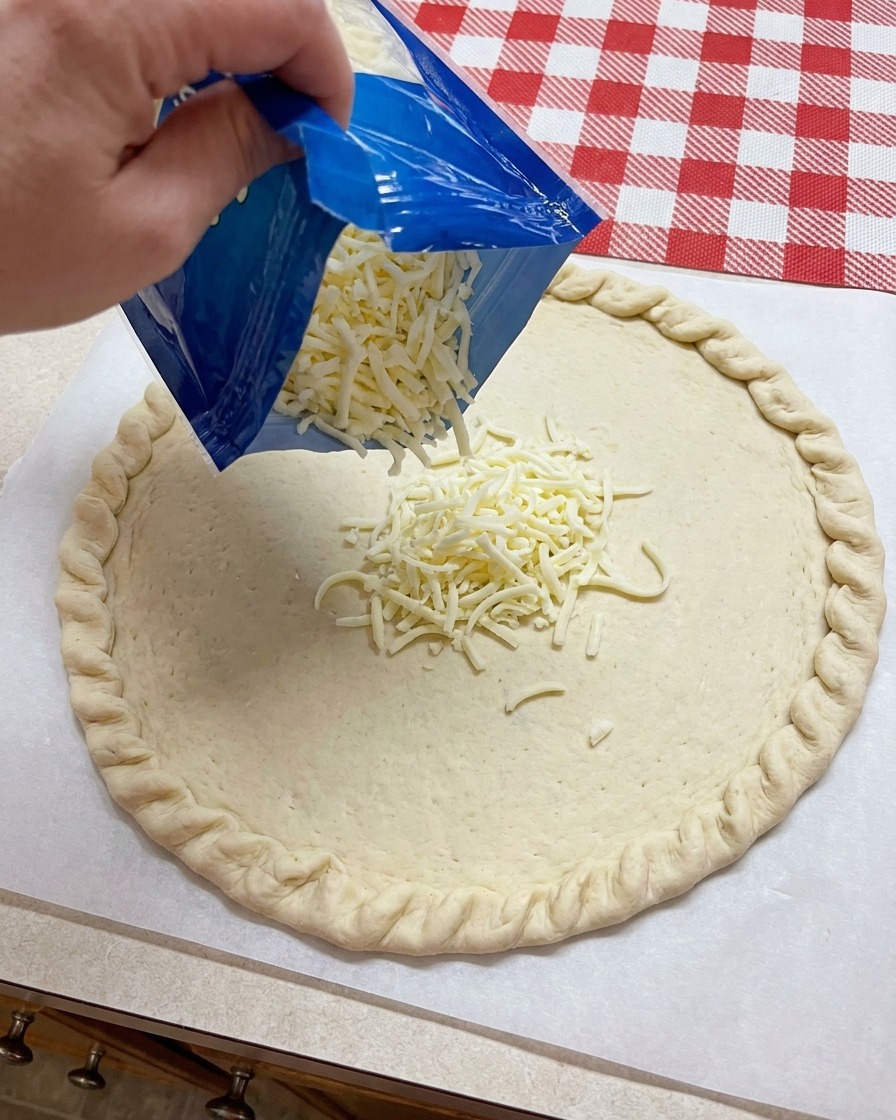Queso mozzarella rallado siendo vertido desde una bolsa azul sobre el centro de una base de masa de pizza moldeada.