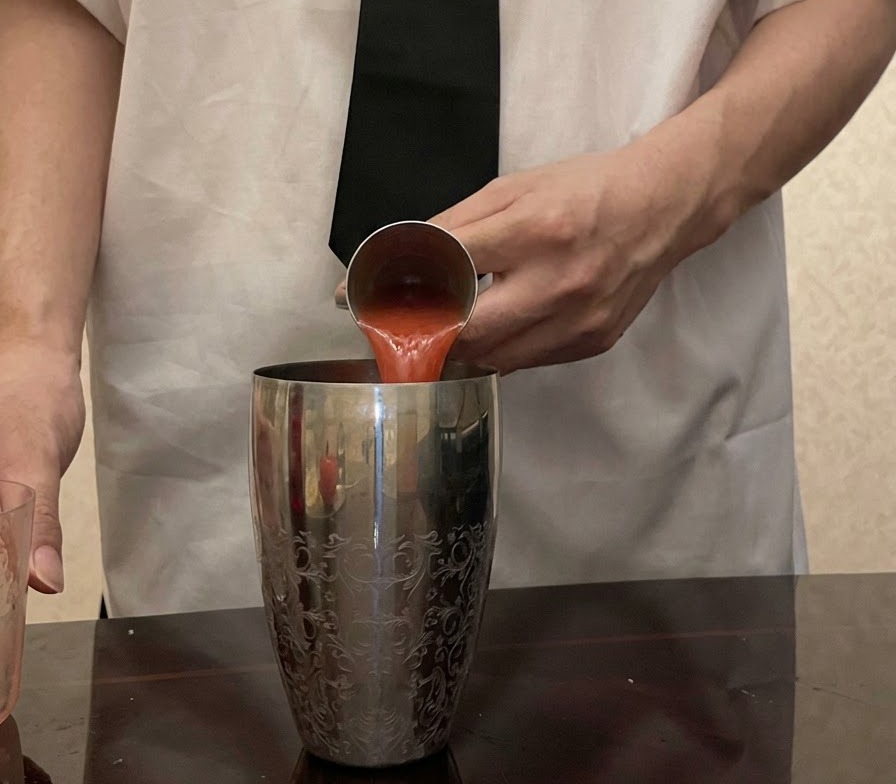 Jugo de tomate rojo fresco siendo vertido desde un medidor de metal en una coctelera de plata con patrones.