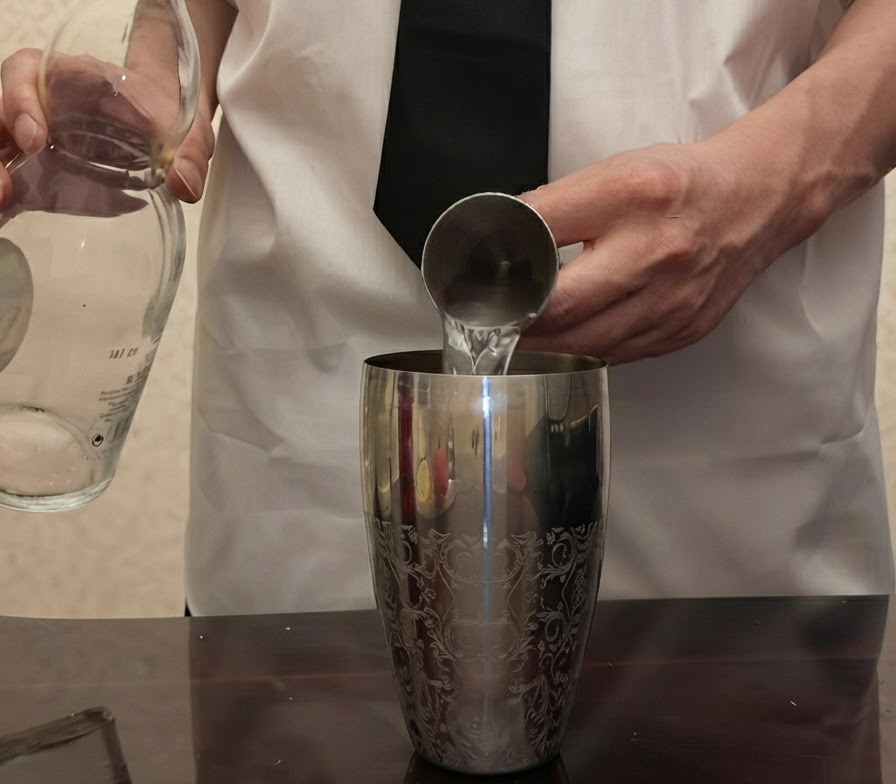 Tequila transparente siendo vertido desde una botella de vidrio en un medidor de metal sostenido sobre una coctelera de plata.