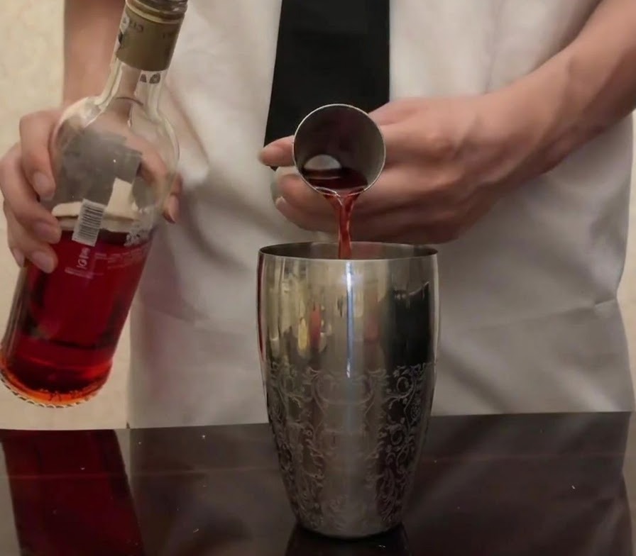 Campari rojo brillante siendo vertido desde su botella en un medidor de metal sobre una coctelera de plata.