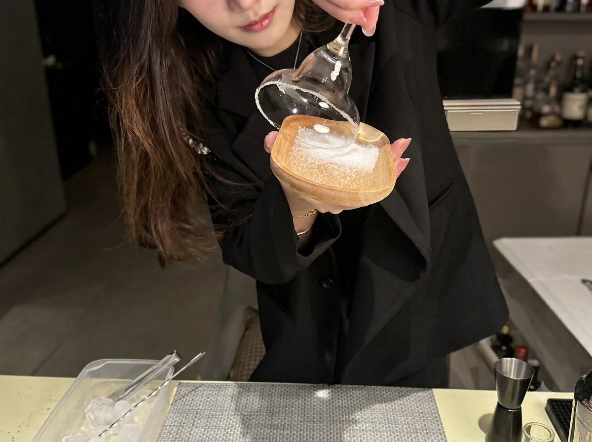 El borde de una copa de cóctel siendo presionado sobre una capa de sal marina en un pequeño plato de madera.