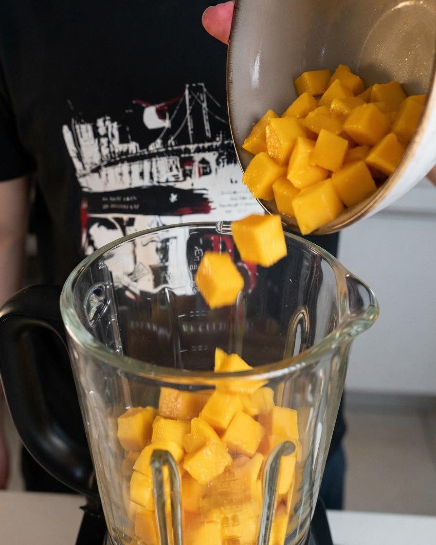 Cubos de mango amarillo recién cortados siendo vertidos desde un bol hacia el vaso transparente de una licuadora.