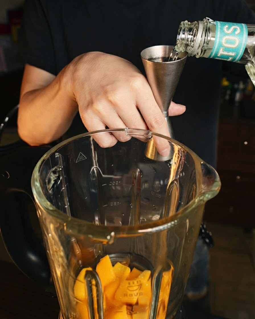 Licor transparente siendo vertido desde un jigger de acero inoxidable en el vaso de la licuadora sobre trozos de mango fresco.
