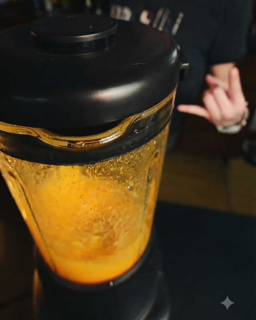Una licuadora funcionando con una mezcla de mango de color naranja brillante en su interior, con una mano haciendo un gesto al fondo.