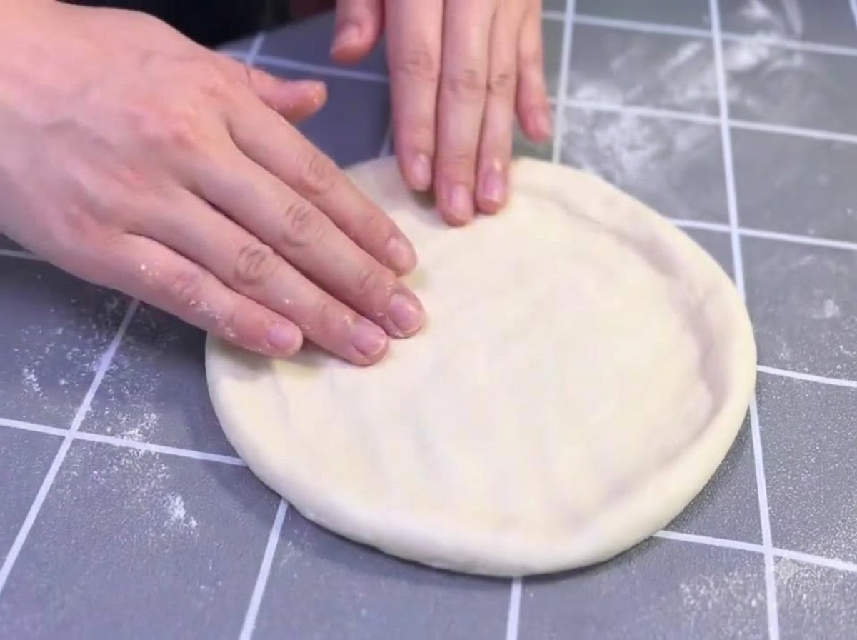 Manos estirando y dando forma suavemente a un círculo de masa de pizza engrasada sobre una superficie cuadriculada gris.