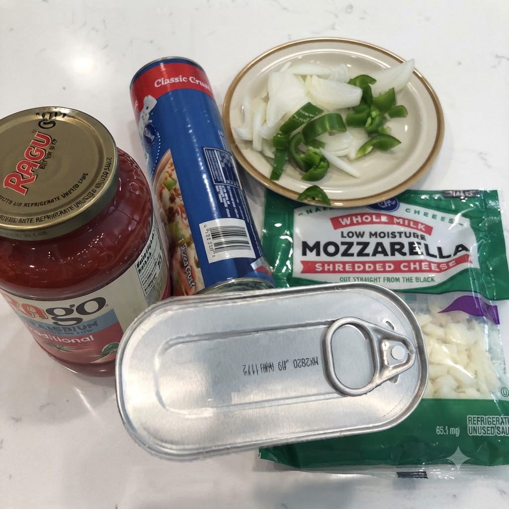 Una lata de anchoas, un tarro de salsa marinara, masa de pizza en lata, mozzarella rallada y cebollas y pimientos cortados en una encimera blanca.