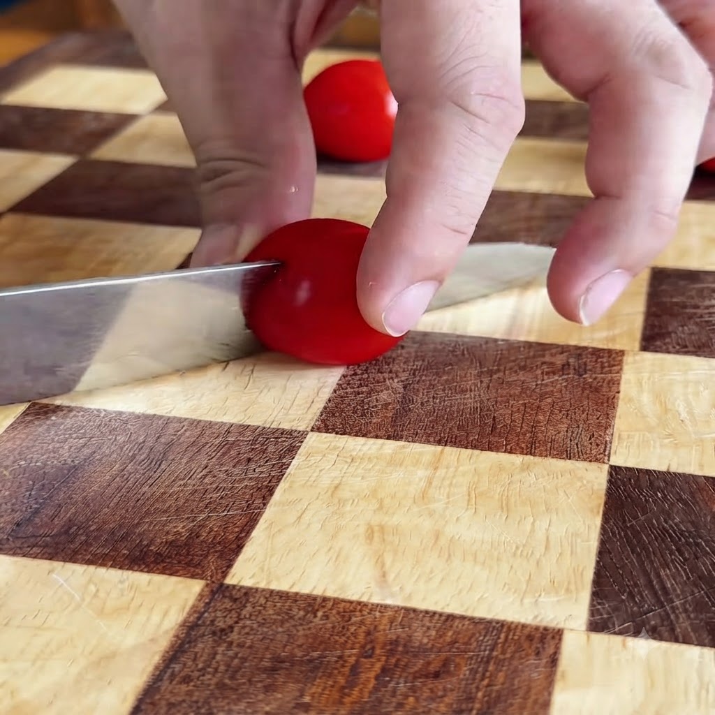 Manos cortando un tomate cherry rojo por la mitad con un cuchillo sobre una tabla de cortar a cuadros.