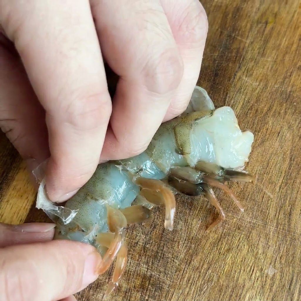 Dedos retirando la cáscara translúcida de un camarón crudo sobre una superficie de madera.