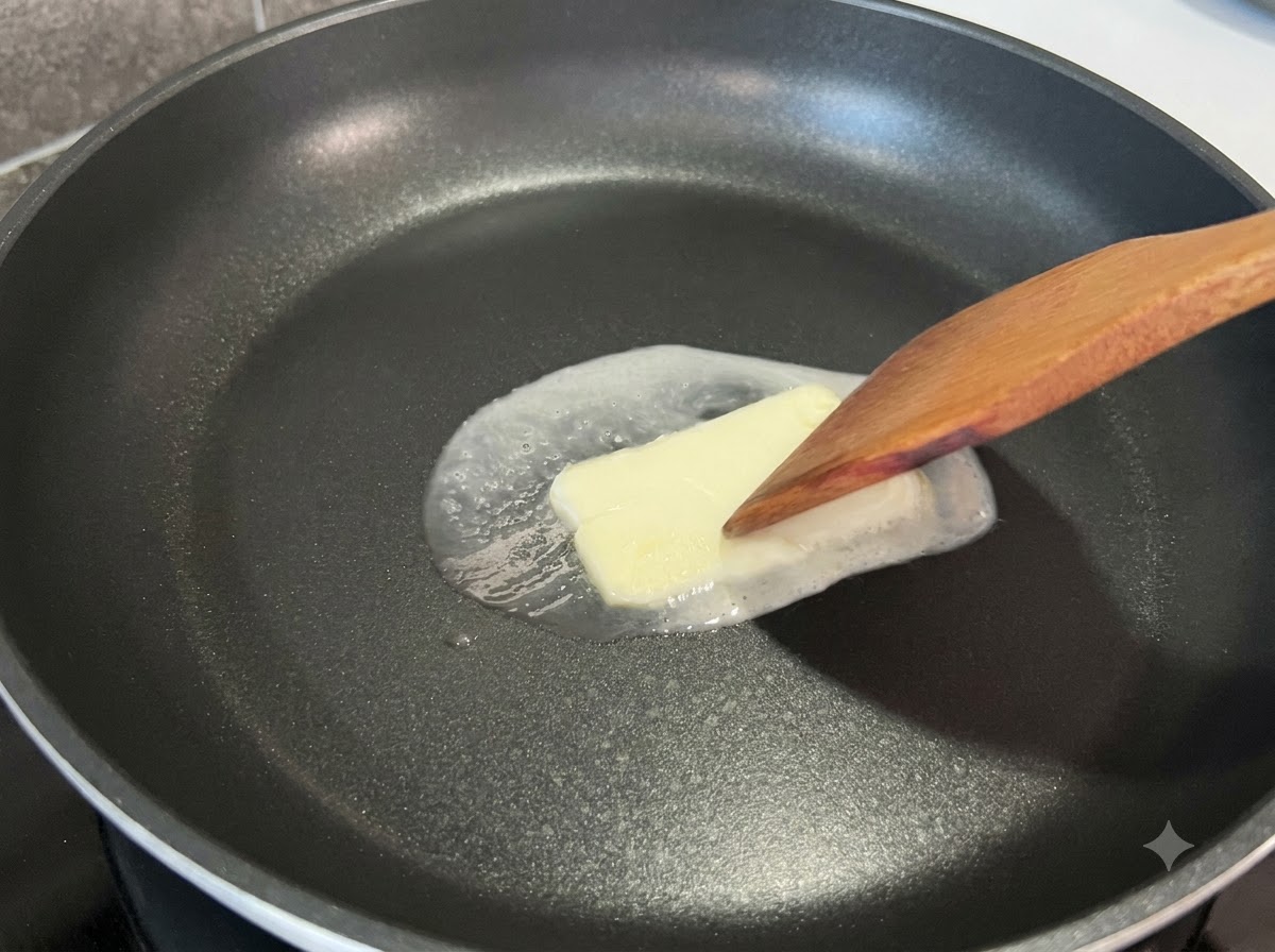 Una espátula de madera presionando ligeramente un trozo cuadrado de mantequilla que se derrite en una sartén negra.