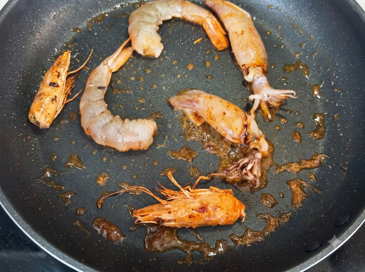 Camarones, cabezas de camarón enteras y trozos de calamar friéndose en una sartén, sazonados con motas oscuras de pimienta negra.